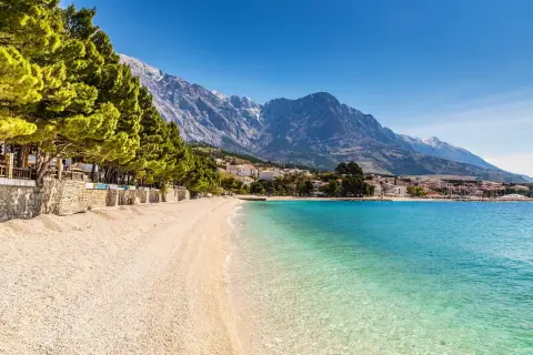 En vacker strand med turkost vatten och en bergskedja i bakgrunden. En promenadstråk kantas av träd och stenmurar, perfekt för en avkopplande stund vid makarska strandpromenad.
