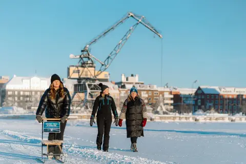 Tre kvinnor njuter av en vinterdag och utforskar Luleå på isen, med en stor kran och snötäckta byggnader i bakgrunden.