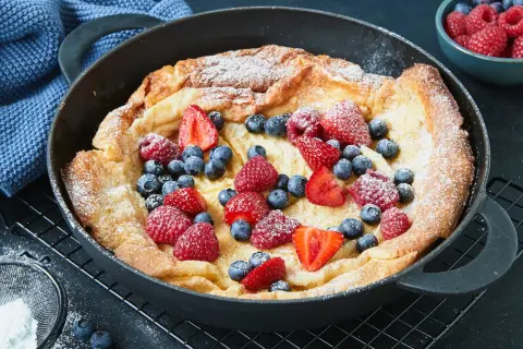 Ein goldbrauner Dutch Baby mit Puderzucker und frischen Beeren wie Erdbeeren, Himbeeren und Blaubeeren in einer Gusseisenpfanne.