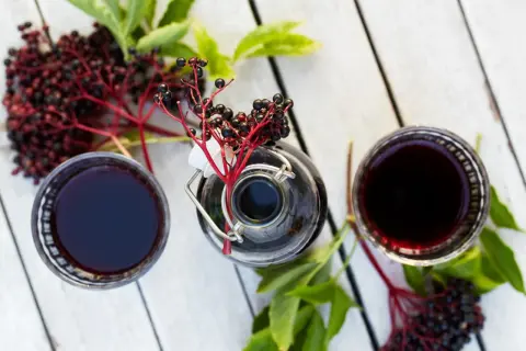 Sok z czarnego bzu ile pić? Dwa kieliszki ciemnego napoju, butelka i gałązki z owocami na białym blacie.