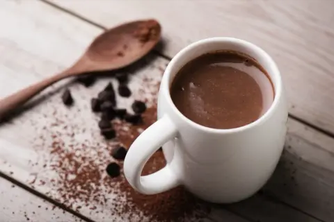 Tazza di cioccolata calda con cacao amaro, gocce di cioccolato e un cucchiaio di legno su un tavolo di legno.