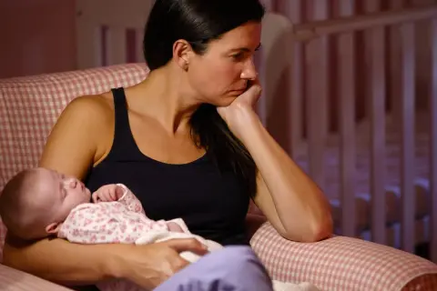Eine Frau sitzt nachdenklich mit ihrem Baby im Arm. Die Szene deutet auf postnatale Depression hin, eine schwere Zeit für viele Mütter.