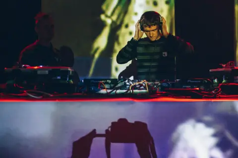 DJ w słuchawkach przy konsoli Pioneer DJ, w tle inny DJ i projekcja.