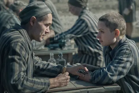Två fångar i randiga uniformer möts vid ett bord. Mannen håller en nål, redo att tatuera kvinnan. Scen från filmen "Tatueraren i Auschwitz".