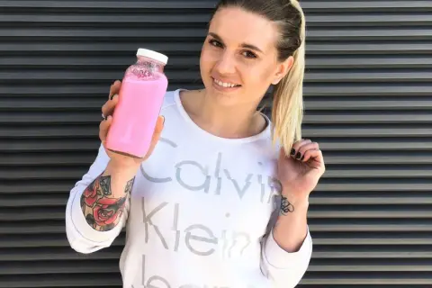 Uśmiechnięta kobieta z tatuażami trzyma różowy shake proteinowy zamiast posiłku.