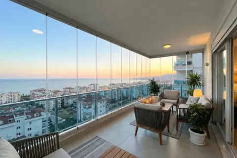 Terraza moderna con cortinas de cristal, muebles de exterior y vistas al mar al atardecer.