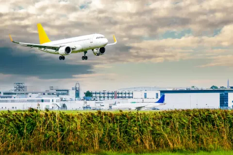 Ein Linienflugzeug mit gelbem Leitwerk nähert sich dem Flughafen. Die Linienflug Geschwindigkeit ist beeindruckend, während es über das Schilf und die Gebäude fliegt.