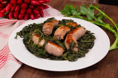Salsiccia e friarielli, un piatto tradizionale napoletano, servito su un piatto bianco con peperoncini rossi sullo sfondo.