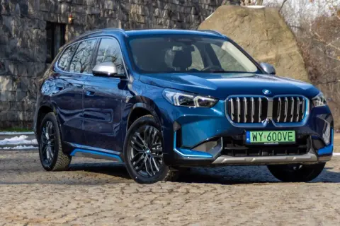 Błękitny BMW X1 elektryk z niebieskimi akcentami na progu, zaparkowany na kamienistej nawierzchni.