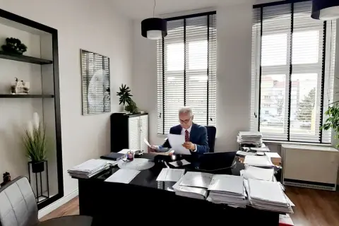 Mężczyzna w garniturze siedzi przy biurku w gabinecie, przeglądając dokumenty.