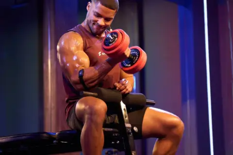 Mann mit Hantel beim Fitness-Training. Schweißglänzende Muskeln, konzentrierter Blick.