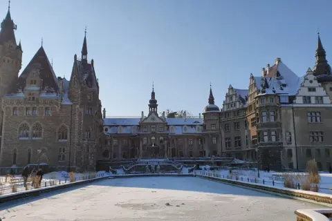 Zamek Moszna w zimowej scenerii, z zamarzniętym stawem i ośnieżonymi dachami. Bajkowa budowla prezentuje się majestatycznie.