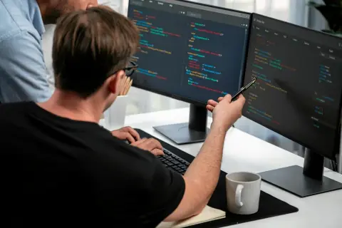 Dwóch programistów analizuje kod na monitorach. To świetny przykład, jak wygląda praca w IT dla początkujących – nauka i współpraca.