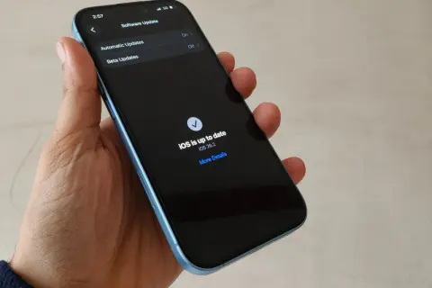 Dłoń trzyma iPhone'a z ekranem pokazującym "iOS is up to date iOS 26.2".