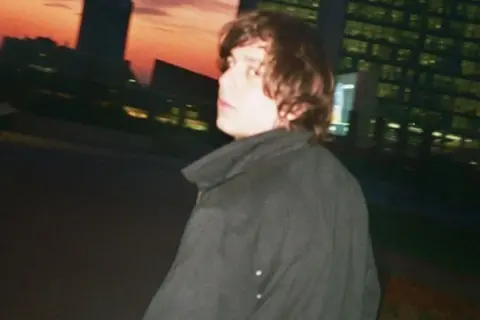 Francesco De Leo, con i capelli castani mossi, indossa un cappotto scuro. Sullo sfondo, un tramonto arancione illumina i profili degli edifici urbani.