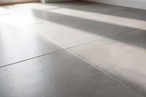Lumière du soleil traversant un carrelage gris, créant des ombres douces. On entendrait presque le carrelage qui sonne creux sous les pas.