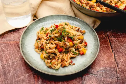 Ein Teller mit kritharaki salat mit hackfleisch, garniert mit frischen Kräutern. Bunte Paprikawürfel und Hackfleisch sind unter die Nudeln gemischt.