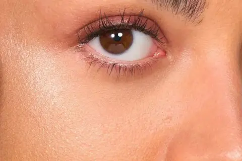Primer rostro con brocha negra aplicando producto no comedogénico. Piel radiante y ojos marrones con sombras rosadas.