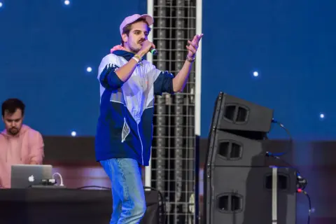 Artysta na scenie, jeden z najpopularniejszych polskich wykonawców pop, śpiewa do mikrofonu. W tle DJ przy laptopie.