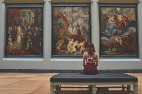 Une femme assise sur un banc, contemplant trois tableaux baroques. L'analyse photo révèle une scène de musée immersive.