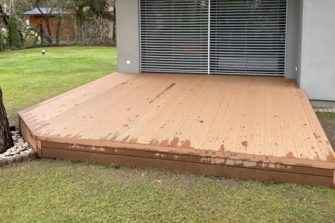 Nowa deska kompozytowa na kostce brukowej, otoczona kamieniami, tworzy nowoczesną przestrzeń przy domu.