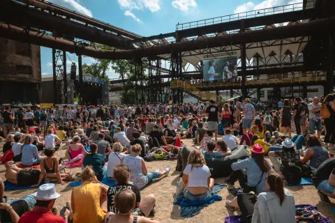 Tłum ludzi na festiwalu muzycznym, siedzących na ziemi i słuchających koncertu. W tle widać scenę i wielki ekran z występującymi artystami. To część owf lineup.