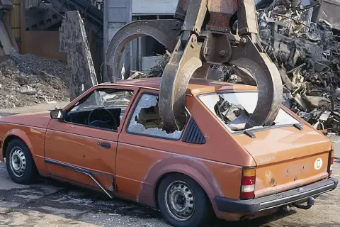 Pomarańczowy samochód z pękniętymi szybami jest podnoszony przez chwytak. Zastanawiasz się, ile kosztuje złomowanie auta?