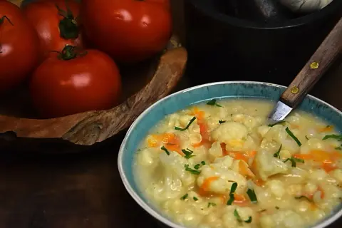 Pyszna zupa kalafiorowa z makaronem, ozdobiona natką pietruszki. Obok miski leżą świeże pomidory.