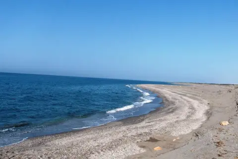 Mar azul y cielo despejado en **Punta Entinas**. La arena y las conchas forman una curva suave hacia el mar.