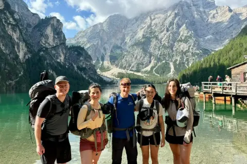 Fem glada vandrare med stora ryggsäckar njuter av en vacker sjö och berg i Italien.