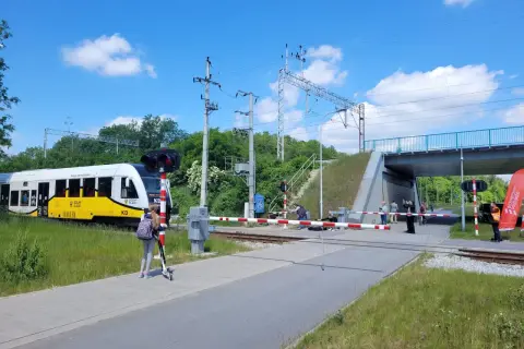 Pociąg KD zbliża się do przejazdu kolejowego. Dziewczynka na hulajnodze czeka na sygnał, by móc kontynuować wicie dojazd PKP.