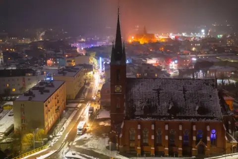 Nocna panorama Kościerzyny, ośnieżona trasa miejska z kościołem w centrum i rozświetlonymi budynkami.