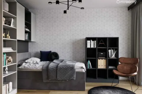 Nowoczesna aranżacja pokoju 10m2: łóżko z szarą pościelą, regały z książkami, czarne meble i brązowy fotel.