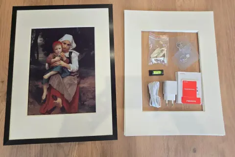 Cadre noir avec une peinture de mère et enfant, à côté d'un cadre blanc contenant des accessoires de montage, comme un niveau à bulle, un chargeur et des adhésifs, rappelant l'effet fresnel d'une œuvre d'art.