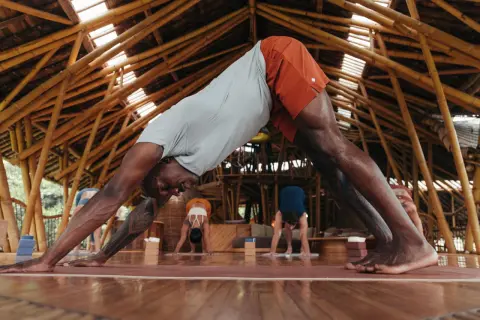 Un homme en posture de chien tête en bas, cherchant à redresser son dos. D'autres pratiquent le yoga dans un studio en bambou.