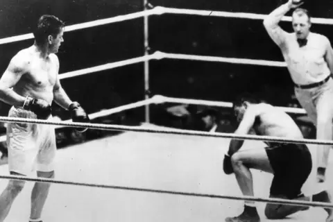 Jack Dempsey vs Gene Tunney in the ring. Dempsey stands, while Tunney is on one knee, with the referee looking on.