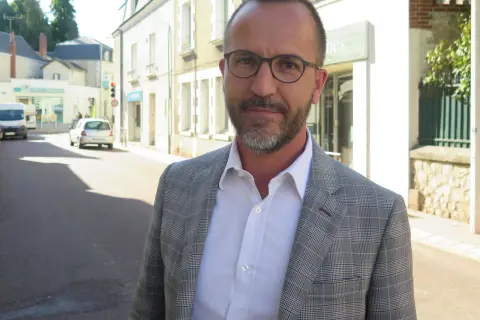 Laurent Raymond, homme politique, porte une veste à carreaux et une chemise blanche, debout dans une rue ensoleillée.