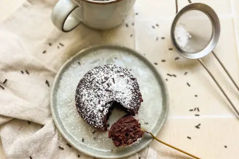 Ein saftiger Schokoladen-Tassenkuchen ohne Ei, bestäubt mit Puderzucker und Schokostreuseln, wartet auf dem Teller.