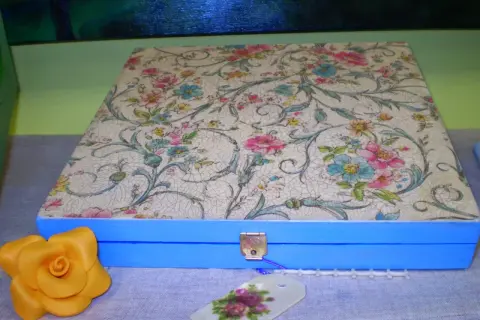 Caja azul con tapa decorada con papel decoupage floral. Un detalle de rosa amarilla y etiqueta floral completan la escena.