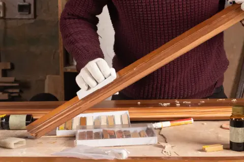 Persona con guantes blancos restaura un mueble de madera, usando un bloque para lijar y kits de retoque.