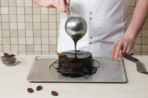 Ein Konditor überzieht einen Kuchen mit glänzender mirror glaze. Schokoladenstückchen liegen daneben.