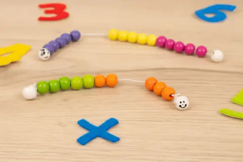 Bunte Perlenketten zum **rechenkette basteln** liegen auf Holz. Zahlen und ein blaues Kreuz sind im Hintergrund.