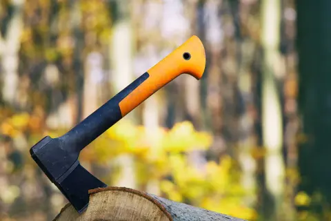 Un hacha con mango naranja y negro clavada en un tronco, lista para el trabajo en el bosque otoñal.