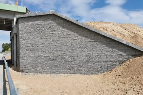Mur oporowy z szarej kostki brukowej, obok nasypu ziemnego i fragmentu wiaduktu.