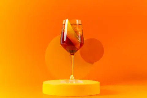 Ein Glas Aperitif mit Orangenscheibe auf gelber Unterlage vor orangefarbenem Hintergrund.