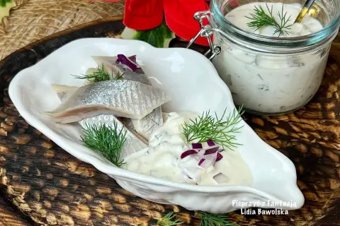 Delikatne śledzie w śmietanie z koperkiem i czerwoną cebulką, podane w białej miseczce. Obok słoiczek z sosem.