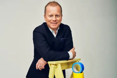 Claes Seldeby, en glad man i kostym, lutar sig mot en gul pall med blå och gul tejp.