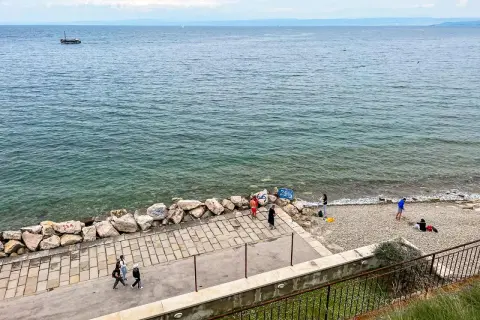 Spacerem po wybrzeżu w Słowenii Piran. Widać łódź na morzu i kilka osób odpoczywających na kamienistej plaży.