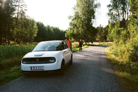 Vit elbil parkerad vid en skogsväg, redo för en bilsemester i Europa. Två personer står vid sidan av vägen.