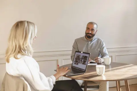Två personer vid ett runt bord, en kvinna med blont hår och en man med skägg. De har en laptop och anteckningsblock, vilket tyder på ett konstruktivt samtal.